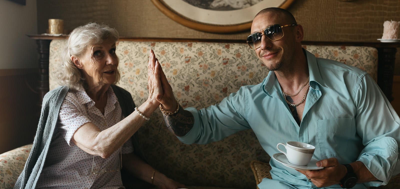 Squareme Loka coffee TV ad - Slovenian celebrity Denis Porčič - Chorchyp sitting on a vintage couch holding a cup of coffee and doing a high five with actress playing a cool grandma
