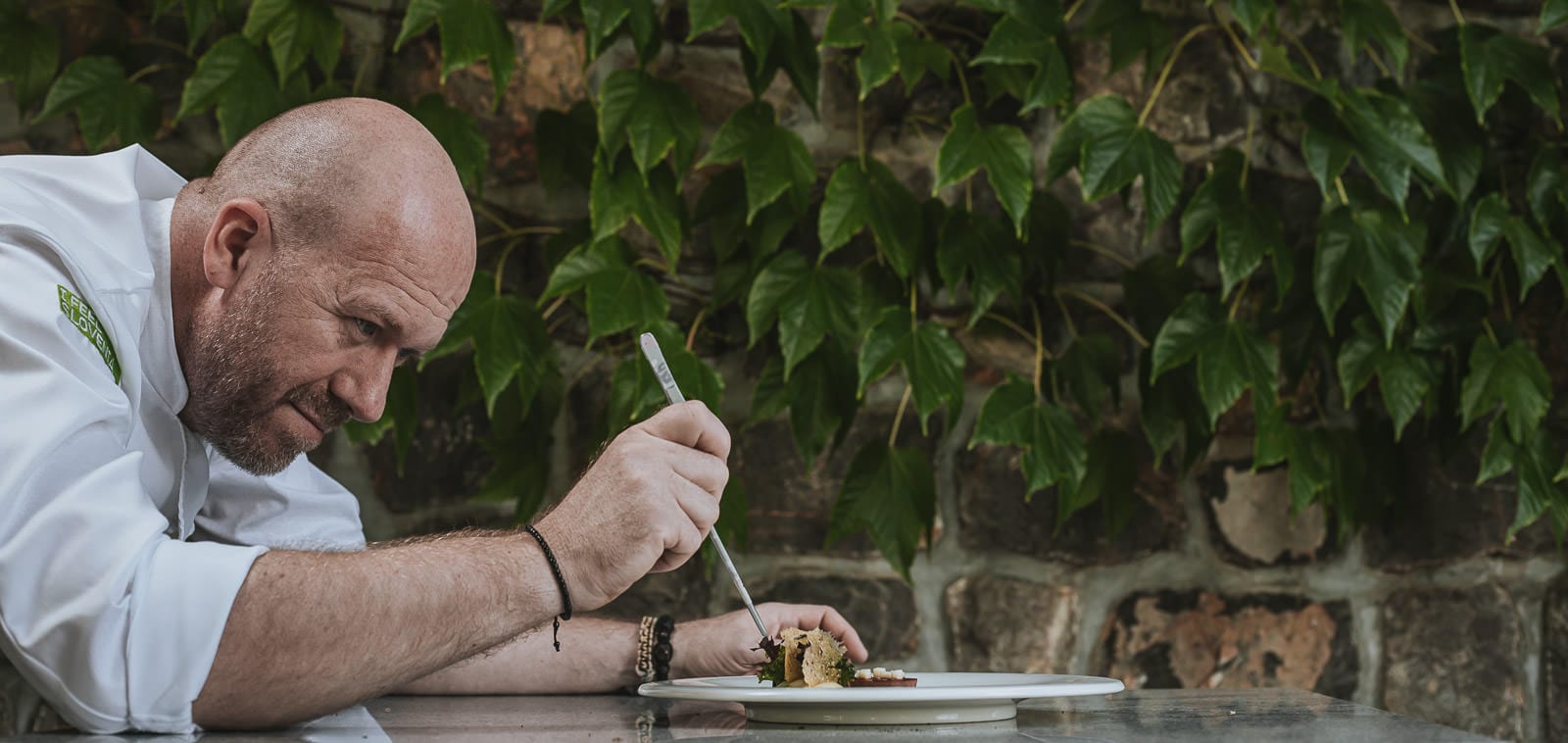 Squareme Mastercard Sous Chef campaign for Michelin star winners in Slovenia - Uroš Štefelin from restaurant Vila Podvin is carefully decorating a plate with plating tweezers in front of an ivy covered brick wall